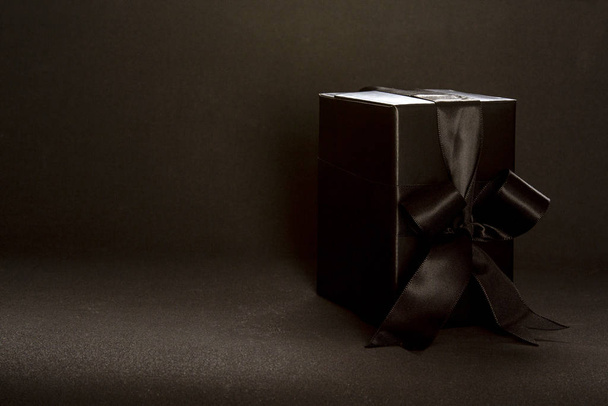Black gift box with black ribbon bow on dark background, copy space. Valentine's day, birthday romantic concept, greeting card. - 写真・画像