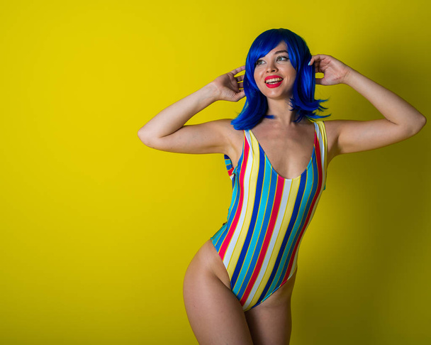 Beautiful woman in a striped swimsuit in a blue wig posing on a yellow background. Portrait of a glamorous girl. - Photo, image