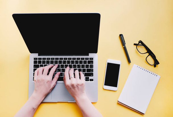 Плоская укладка рабочего стола с женскими руками, печатающими на ноутбуке со смартфоном, бумагой для заметок и очками на ярко-желтом фоне, концепция бизнеса и образа жизни
 - Фото, изображение