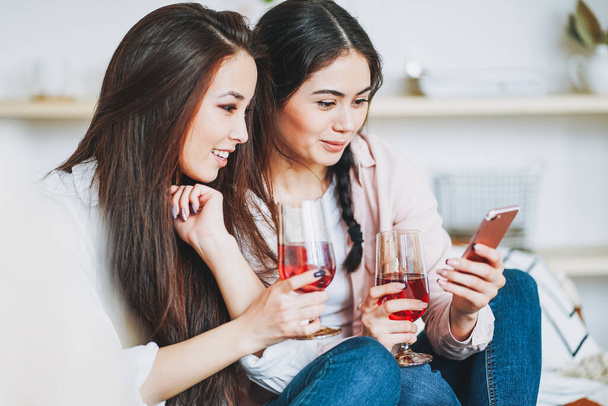 Young carefree brunette girls friends in casual with glasses of wine using mobile and having fun together on home party - Photo, Image
