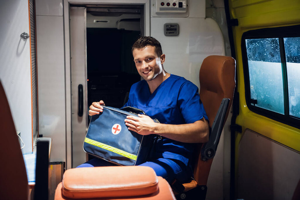 Санитар в медицинской форме сидит в машине скорой помощи и упаковывает медицинскую сумку.
 - Фото, изображение