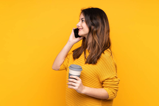 Junge kaukasische Frau isoliert auf gelbem Hintergrund mit Kaffee zum Mitnehmen und einem Handy - Foto, Bild