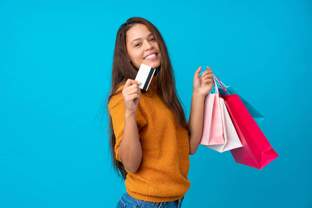 Jeune fille brésilienne sur fond bleu isolé tenant des sacs à provisions et une carte de crédit - Photo, image