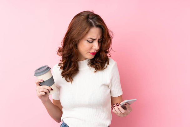 Jonge Russische vrouw over geïsoleerde roze achtergrond houden koffie mee te nemen en een mobiele - Foto, afbeelding