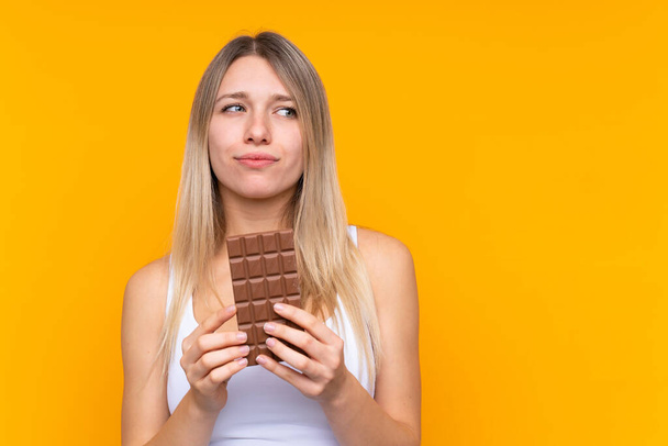 Jonge blonde vrouw over geïsoleerde blauwe achtergrond nemen van een chocolade tablet en met twijfels - Foto, afbeelding