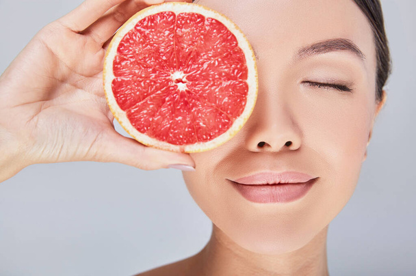 Close-up, a slice of grapefruit near the face of an Asian woman. - Foto, afbeelding