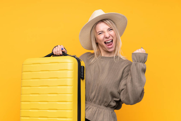 Junge blonde Frau über isoliertem gelben Hintergrund im Urlaub mit Reisekoffer und Hut - Foto, Bild