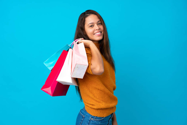 Young Brazilian girl over isolated blue background holding shopping bags and smiling - Fotoğraf, Görsel