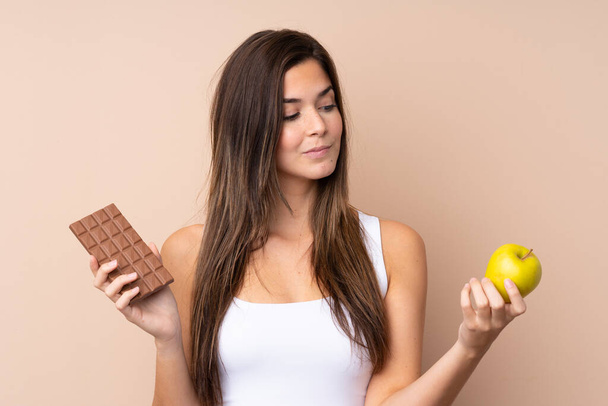 Teenager girl over isolated background having doubts while taking a chocolate tablet in one hand and an apple in the other - Фото, изображение