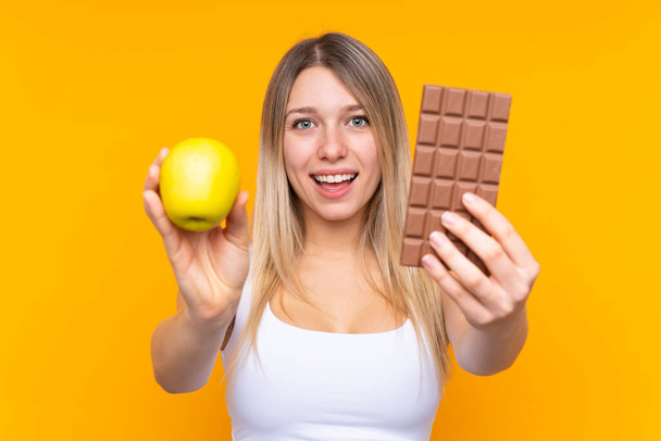 Junge blonde Frau vor isoliertem blauen Hintergrund, die in der einen Hand eine Schokotablette und in der anderen einen Apfel nimmt - Foto, Bild