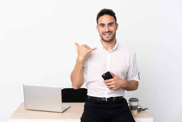 Joven hombre de negocios con un teléfono móvil en un lugar de trabajo haciendo un gesto de teléfono
 - Foto, Imagen