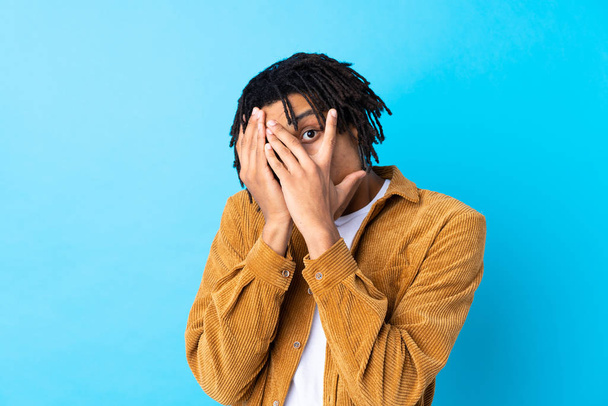 junger afrikanisch-amerikanischer Mann mit Cordjacke über isoliertem blauem Hintergrund, der Augen bedeckt und durch Finger schaut - Foto, Bild