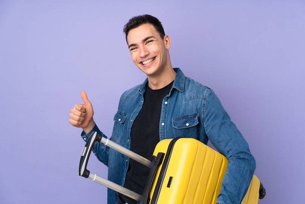młody biały przystojny mężczyzna odizolowany na fioletowy tle na wakacjach z podróżną walizką i z kciukiem w górze - Zdjęcie, obraz