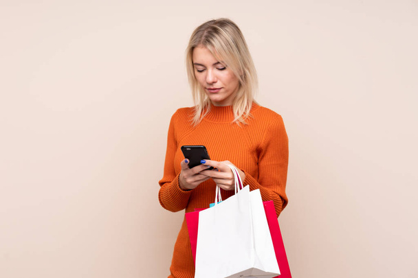 Young blonde woman over isolated background holding shopping bags and writing a message with her cell phone to a friend - Photo, Image