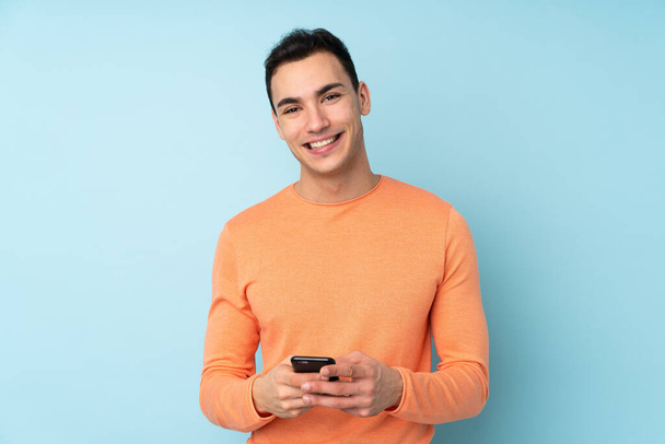 Young caucasian handsome man isolated on blue background sending a message with the mobile - Photo, Image