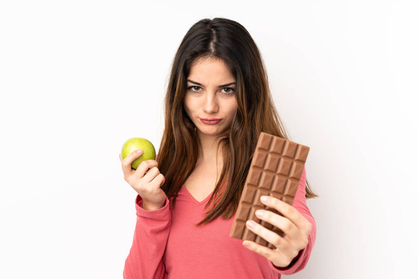 Junge kaukasische Frau isoliert auf rosa Hintergrund mit einer Schokoladentablette in der einen Hand und einem Apfel in der anderen - Foto, Bild