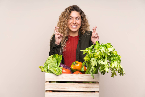 Agricultor con verduras recién recogidas en una caja aislada sobre fondo beige con los dedos cruzados - Foto, Imagen
