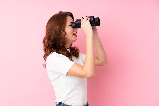 jong russisch vrouw over geïsoleerde roze achtergrond met zwart verrekijker - Foto, afbeelding