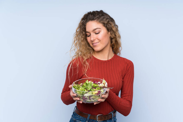 Young blonde woman with curly hair holding a salad over isolated wall - Fotografie, Obrázek