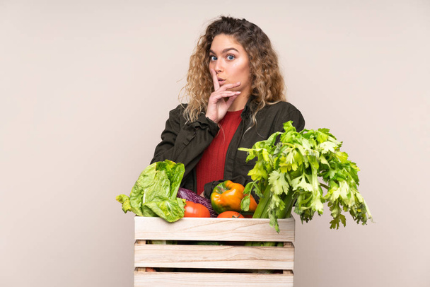 Agricultor con verduras recién recogidas en una caja aislada sobre fondo beige haciendo gesto de silencio - Foto, Imagen