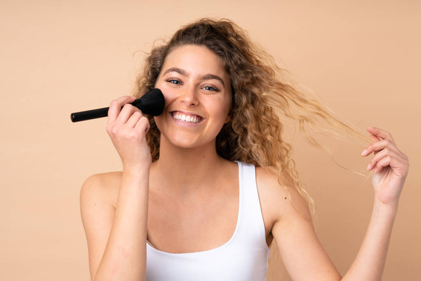 Young blonde woman with curly hair isolated on beige background holding makeup brush - Photo, Image