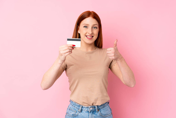 Young redhead woman over isolated pink background holding a credit card - Fotoğraf, Görsel