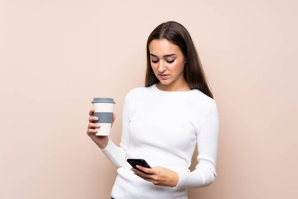 Νεαρή γυναίκα πάνω από απομονωμένη φόντο κρατώντας καφέ για να πάρει μακριά και ένα κινητό - Φωτογραφία, εικόνα