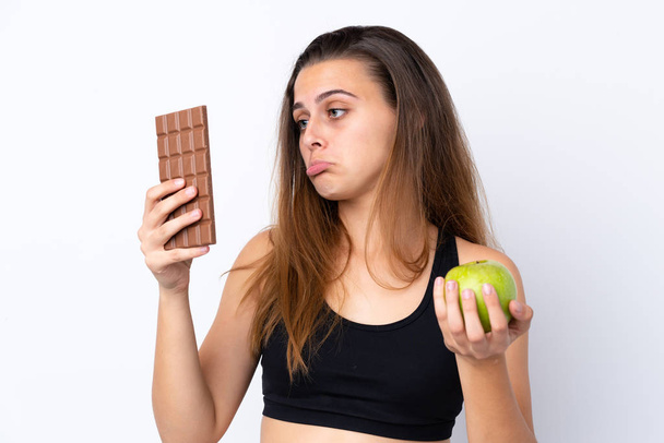 Дівчина-підліток над ізольованим білим тлом має сумніви, приймаючи шоколадну таблетку в одній руці і яблуко в іншій
 - Фото, зображення