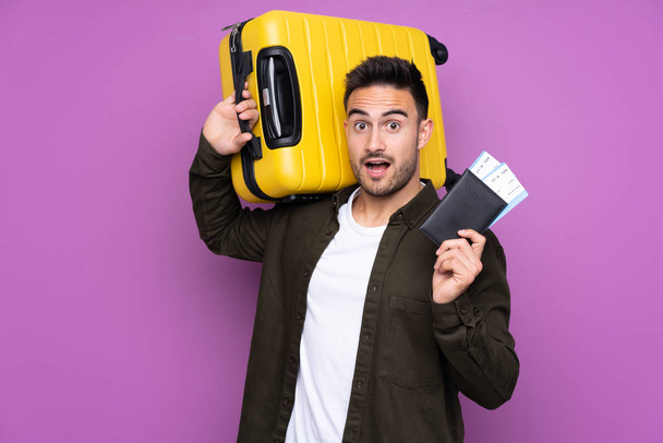 Joven hombre guapo sobre fondo púrpura aislado en vacaciones con maleta y pasaporte y sorprendido
 - Foto, Imagen
