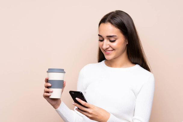 Junge Frau über isolierten Hintergrund mit Kaffee zum Mitnehmen und einem Handy - Foto, Bild