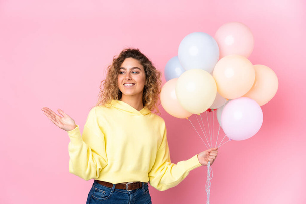 Jovem loira com cabelos cacheados pegando muitos balões isolados no fundo rosa estendendo as mãos para o lado para convidar para vir - Foto, Imagem