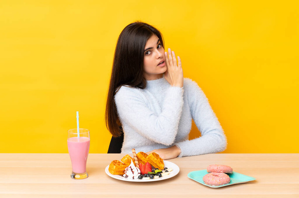 Young woman eating waffles and milkshake in a table over isolated yellow background whispering something - Fotoğraf, Görsel