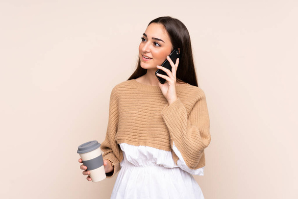 jonge Kaukasische vrouw geïsoleerd op beige achtergrond houden koffie mee te nemen en een mobiele - Foto, afbeelding