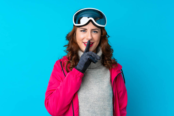 Skiër vrouw met snowboard bril over geïsoleerde blauwe muur doet stilte gebaar - Foto, afbeelding