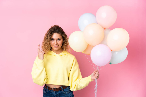 Young blonde woman with curly hair catching many balloons isolated on pink background pointing with the index finger a great idea - Photo, Image