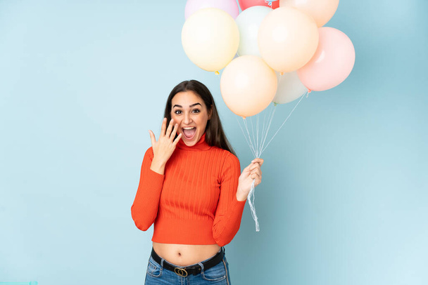 Young woman catching many balloons isolated on blue background with surprise facial expression - 写真・画像