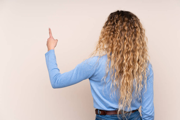 Junge blonde Frau mit lockigem Haar trägt einen Rollkragenpullover auf beigem Hintergrund, der mit dem Zeigefinger nach hinten zeigt - Foto, Bild