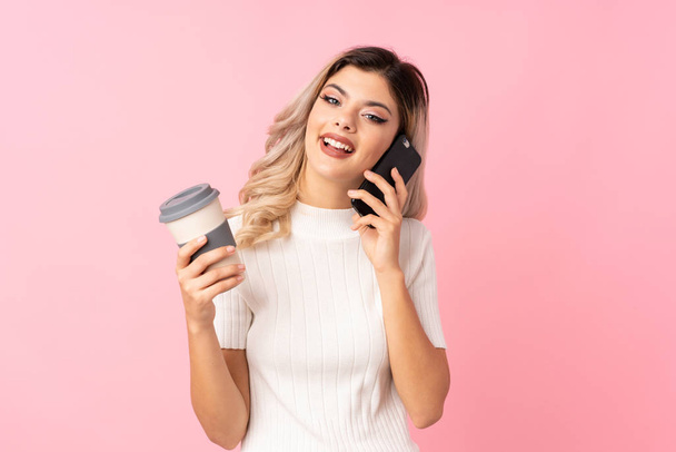 Teenager girl over isolated pink background holding coffee to take away and a mobile - Photo, Image