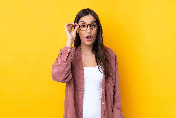 Young brunette woman over isolated yellow background with glasses and surprised - Foto, afbeelding