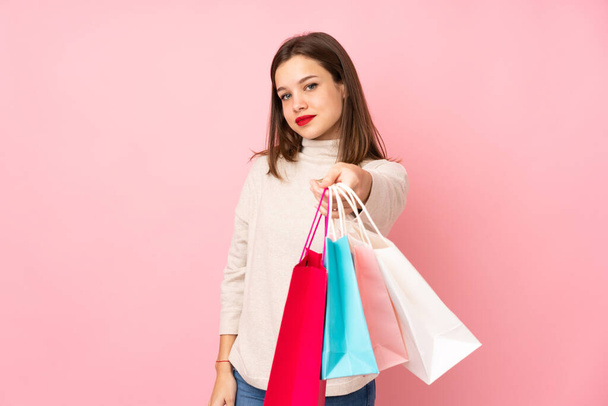Teenager girl isolated on pink background holding shopping bags and giving them to someone - Φωτογραφία, εικόνα