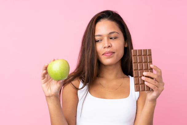 Молодая бразильская девушка на изолированном розовом фоне берет шоколадную таблетку в одной руке и яблоко в другой - Фото, изображение