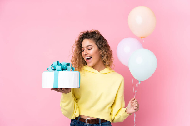 Young blonde woman with curly hair catching many balloons and holding a big cake isolated on pink background - Photo, image