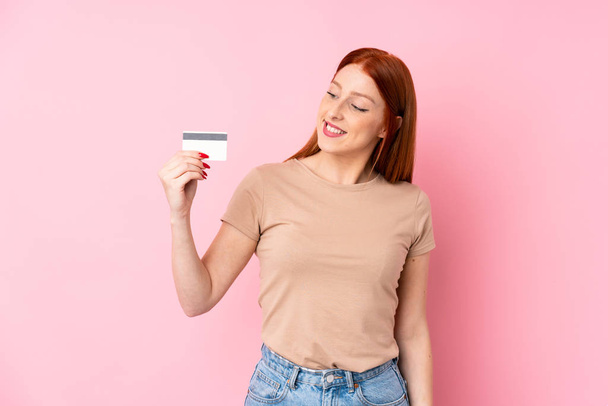 Young redhead woman over isolated pink background holding a credit card - 写真・画像