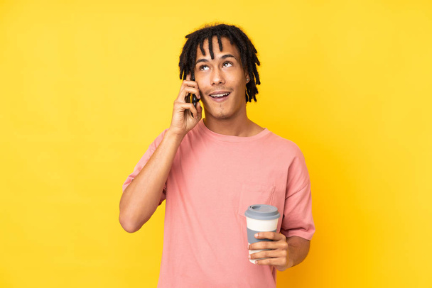 Young african american man isolated on yellow background holding coffee to take away and a mobile - Foto, afbeelding