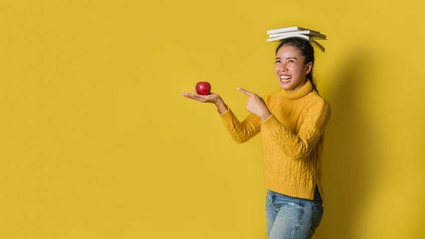Ένα κορίτσι με ένα βιβλίο στο κεφάλι και ένα κόκκινο μήλο στο χέρι, ενώ το άλλο χέρι δείχνει το μήλο σε κίτρινο φόντο. Η έννοια της άσκησης για την καλή υγεία. - Φωτογραφία, εικόνα