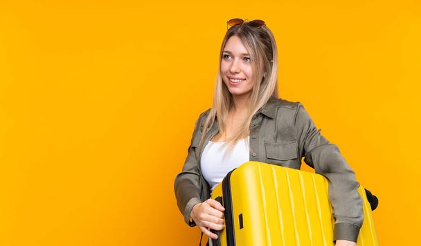 Young blonde woman over isolated yellow background in vacation with travel suitcase and a hat - Photo, image