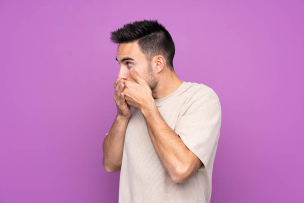 Молодий красивий чоловік над ізольованим фіолетовим тлом, що закриває рот і дивиться в бік
 - Фото, зображення