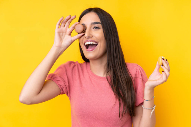 Young brunette woman over isolated yellow background holding colorful French macarons - Fotó, kép