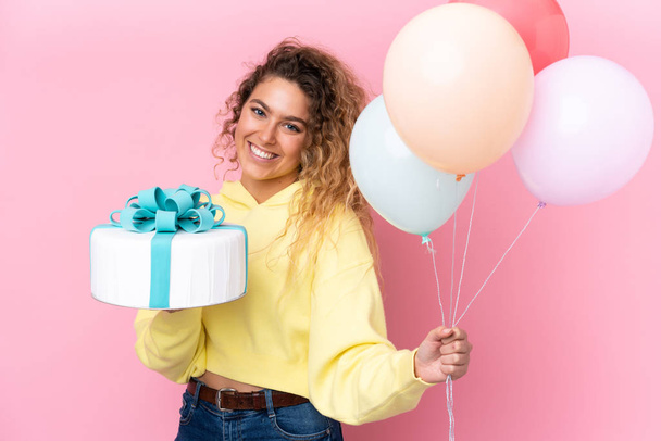 Young blonde woman with curly hair catching many balloons and holding a big cake isolated on pink background - Photo, Image