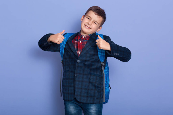 Image of funny playful cute kid being ready to school, raising arms, making gesture, showing sign of approval, super, thumbs up, closing eyes, standing isolated over blue background in studio. - Foto, immagini